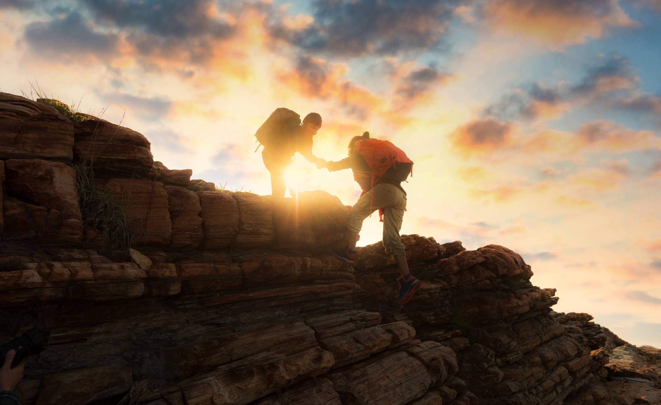 Person helping another up a mountain at sunset.