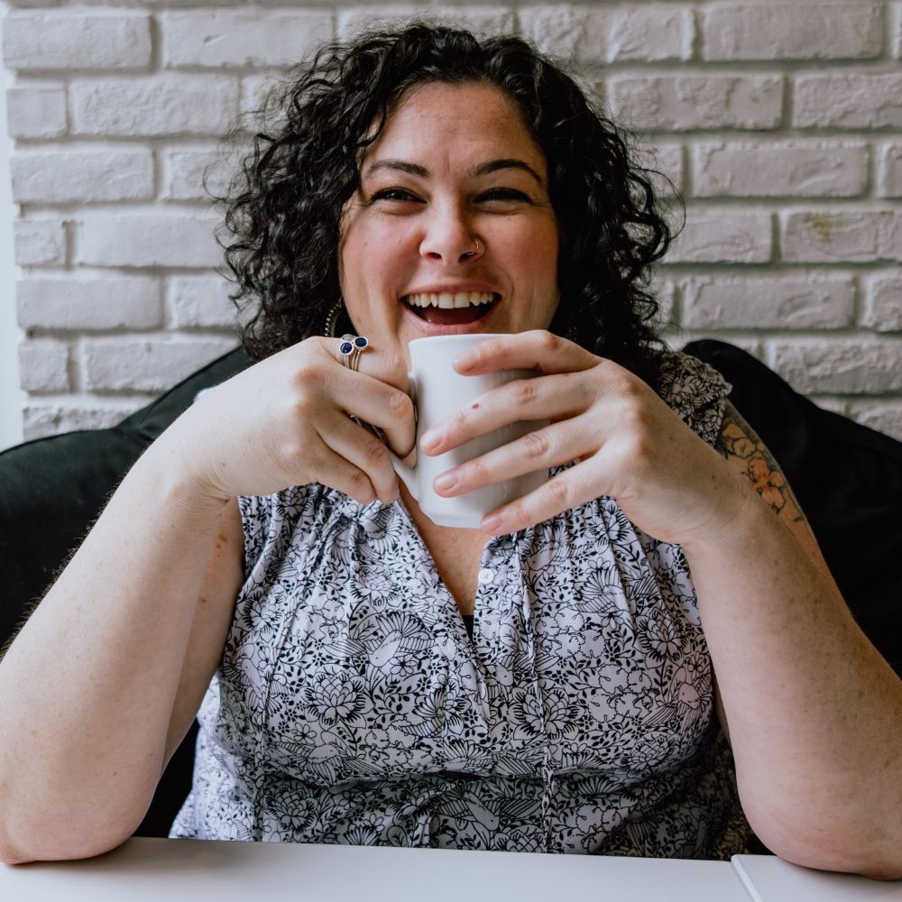 Woman with brown curly hair laughing while holding coffee cup with two hands.
